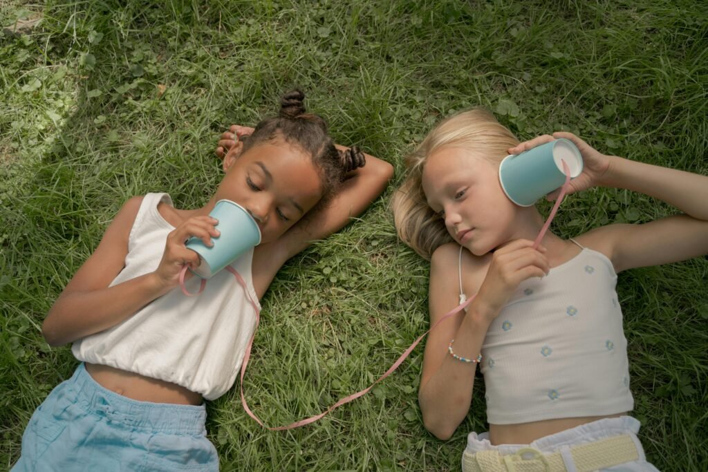 Two young girls lying on grass, using homemade cup phones to communicate and have fun outdoors.