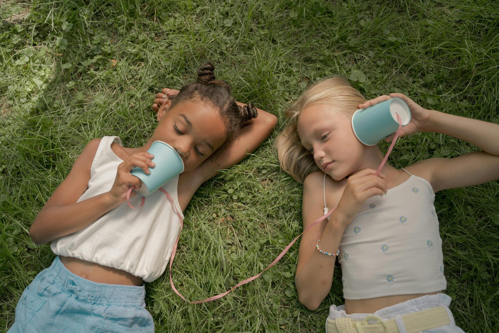 Two young girls lying on grass, using homemade cup phones to communicate and have fun outdoors.